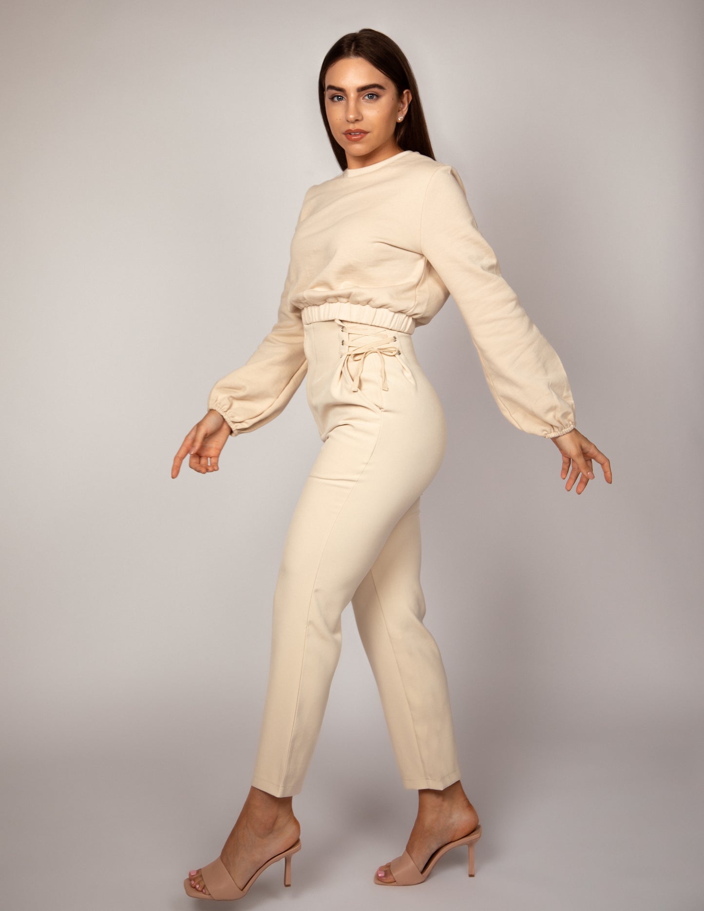 Woman standing sideways with wearing high waisted nude trousers with lace up waist detail and nude jumper with balloon sleeve and elasticated hem.