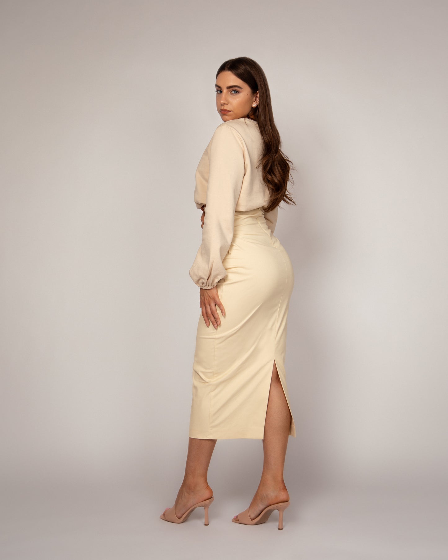 Woman standing with her back to the camera but her face turned to face us over her shoulder.  Wearing a nude jumper with balloon sleeves and a high waisted pencil skirt with lace up waist detail.