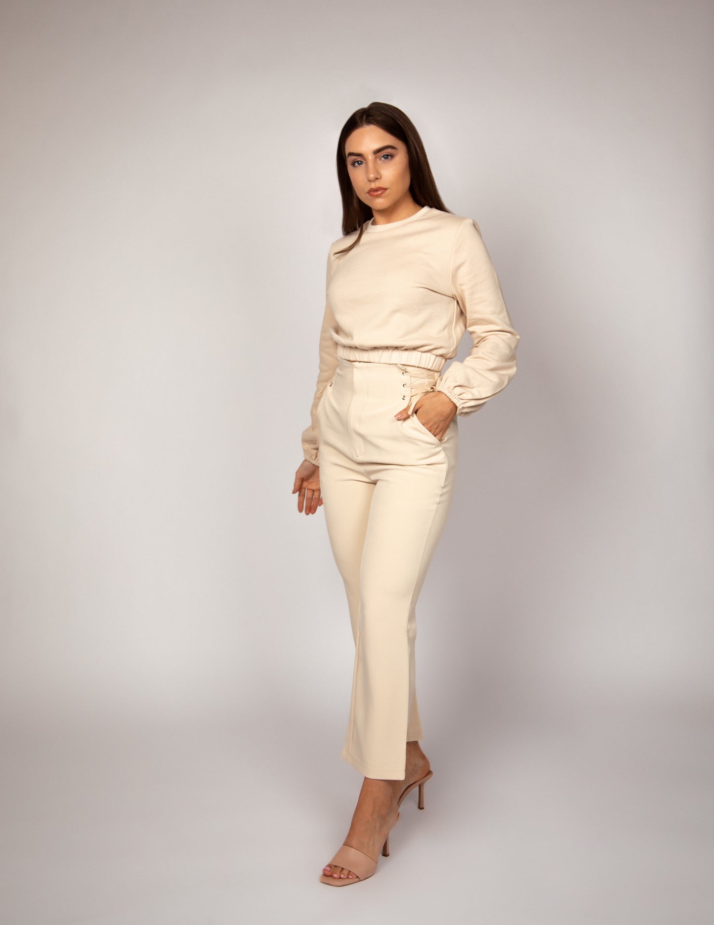 Woman standing with one hand in pocket wearing high waisted nude trousers with lace up waist detail and nude jumper with balloon sleeve and elasticated hem.