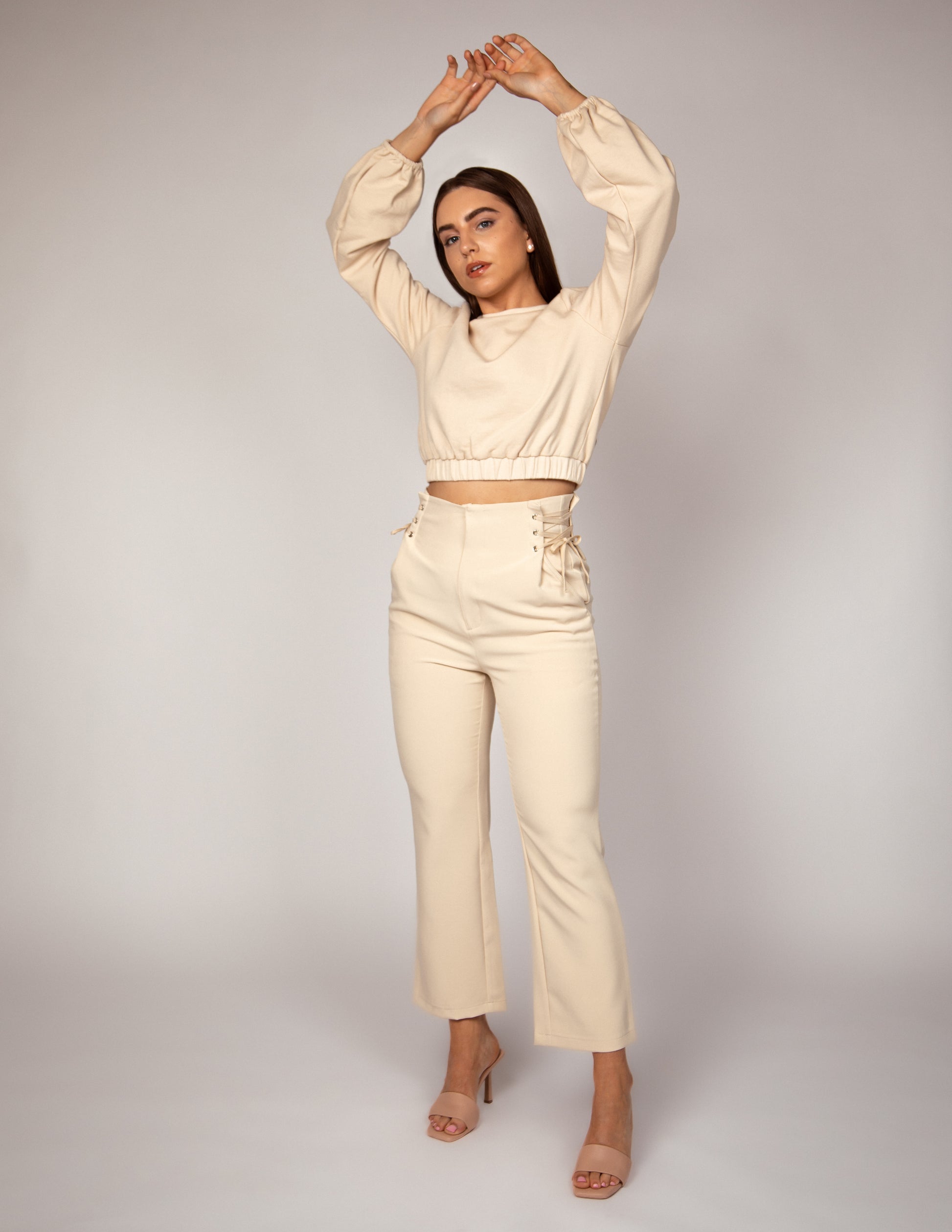 Woman stands with hands raised above her head. She wears high waisted tailored cream/beige/nude trousers that have lace up waist detailing paired with a cream jumper that is slightly cropped and has balloon sleeves.